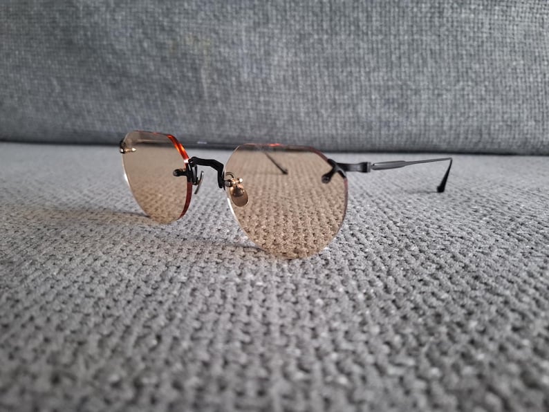 May include: Rimless sunglasses with light brown tinted lenses. The glasses have a black frame and temples, with a small gold-colored accent at the nose bridge. The lenses are a unique octagonal shape.