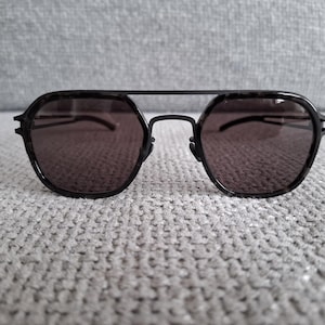 Könnte beinhalten: Schwarze Sonnenbrille mit Doppelsteg und dunklen Gläsern. Die Fassungen haben eine geometrische Form. Die Sonnenbrille liegt auf einer grauen, strukturierten Oberfläche.