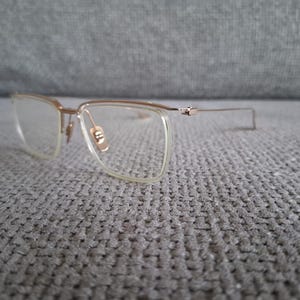 May include: Eyeglasses with a gold-coloured frame and clear lenses. The glasses have a rectangular shape and are on a grey, textured surface. The nose pads are also gold.
