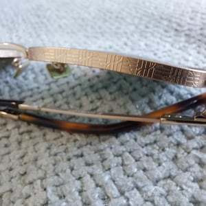Puede incluir: Primer plano de gafas con montura dorada y patillas estampadas. Las lentes est&aacute;n ausentes y las varillas son una mezcla de oro y carey. Las gafas est&aacute;n sobre un fondo azul claro texturizado.