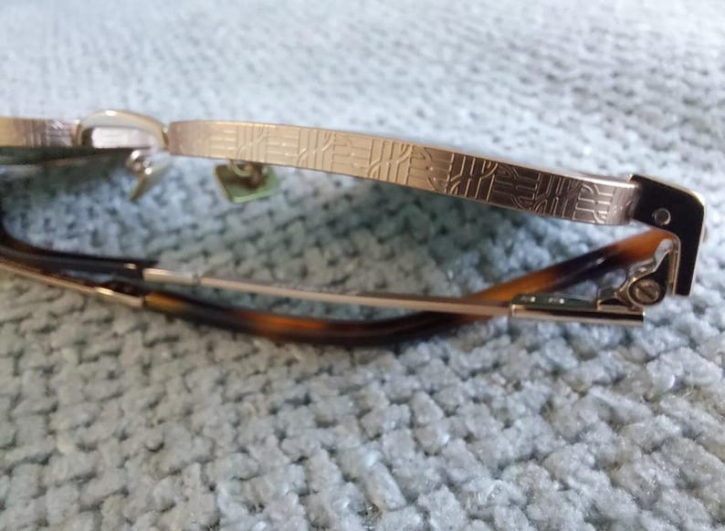 Puede incluir: Primer plano de gafas con montura dorada y patillas estampadas. Las lentes est&aacute;n ausentes y las varillas son una mezcla de oro y carey. Las gafas est&aacute;n sobre un fondo azul claro texturizado.