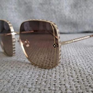 May include: Oversized square sunglasses featuring a gold-tone metal frame with brown gradient lenses. The outer edges of the frames are embellished with small, clear crystals, and a small decorative floral-shaped accent sits on the outer corners.