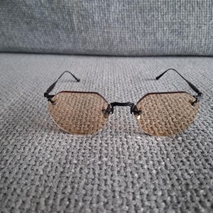 May include: A pair of rimless sunglasses with a black frame and tinted, light brown lenses. The lenses have a unique, geometric shape. The glasses are resting on a gray, textured surface.