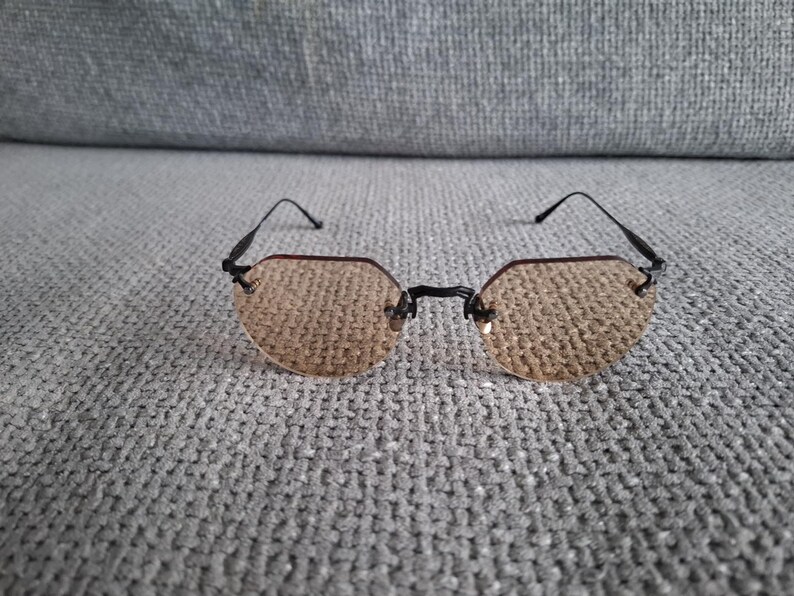 May include: A pair of rimless sunglasses with a black frame and tinted, light brown lenses. The lenses have a unique, geometric shape. The glasses are resting on a gray, textured surface.