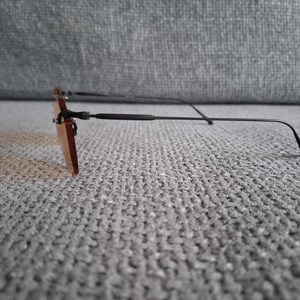 May include: Rimless sunglasses with rectangular, tinted lenses and thin, black metal frames. The temples curve slightly at the ends. The sunglasses are resting on a textured, gray surface.