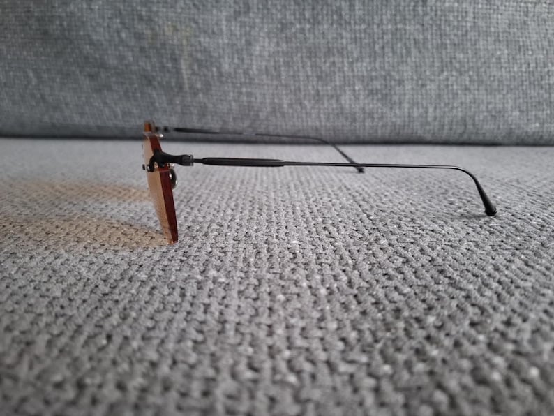 May include: Rimless sunglasses with rectangular, tinted lenses and thin, black metal frames. The temples curve slightly at the ends. The sunglasses are resting on a textured, gray surface.