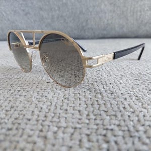 Puede incluir: Gafas de sol con montura dorada, doble puente y lentes oscuras. Las patillas son negras con detalles dorados. Las gafas de sol est&aacute;n sobre una superficie texturizada de color gris claro.