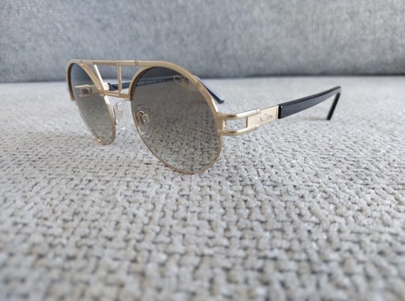 Puede incluir: Gafas de sol con montura dorada, doble puente y lentes oscuras. Las patillas son negras con detalles dorados. Las gafas de sol est&aacute;n sobre una superficie texturizada de color gris claro.