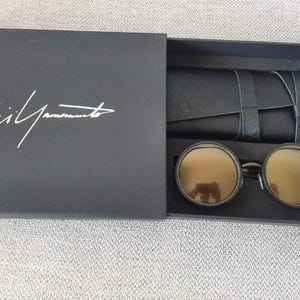 May include: Black box with the white signature of Yohji Yamamoto. Inside, round sunglasses with gold-tinted lenses and a black leather case. The sunglasses have a dark metal frame.