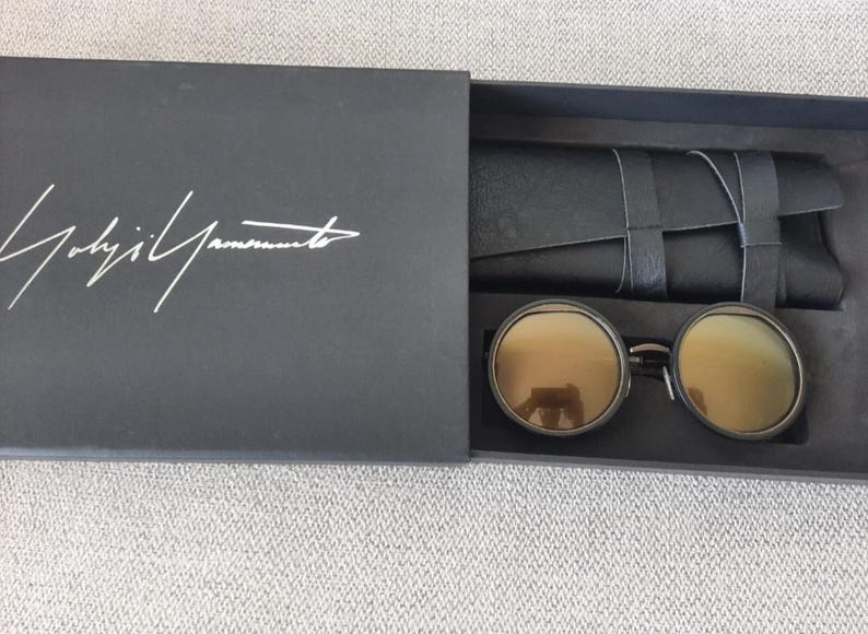 May include: Black box with the white signature of Yohji Yamamoto. Inside, round sunglasses with gold-tinted lenses and a black leather case. The sunglasses have a dark metal frame.