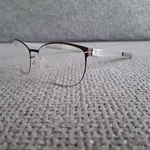 Może przedstawiać: Okulary z ciemnobrązową oprawką i srebrnymi zausznikami. Soczewki są przezroczyste, a okulary spoczywają na szarej, teksturowanej powierzchni. Design jest nowoczesny i minimalistyczny.