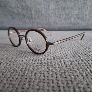 May include: Round-framed eyeglasses with a dark brown rim and temples. The lenses are clear, and the frame has a silver-colored bridge. The glasses rest on a gray textured surface. The word "VISION" is printed on the lens.