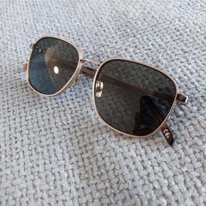 Puede incluir: Gafas de sol cuadradas con montura de metal dorado y lentes oscuras. Las gafas de sol est&aacute;n sobre una superficie texturizada de color gris claro. Las lentes son reflectantes y las patillas tienen un dise&ntilde;o sutil.