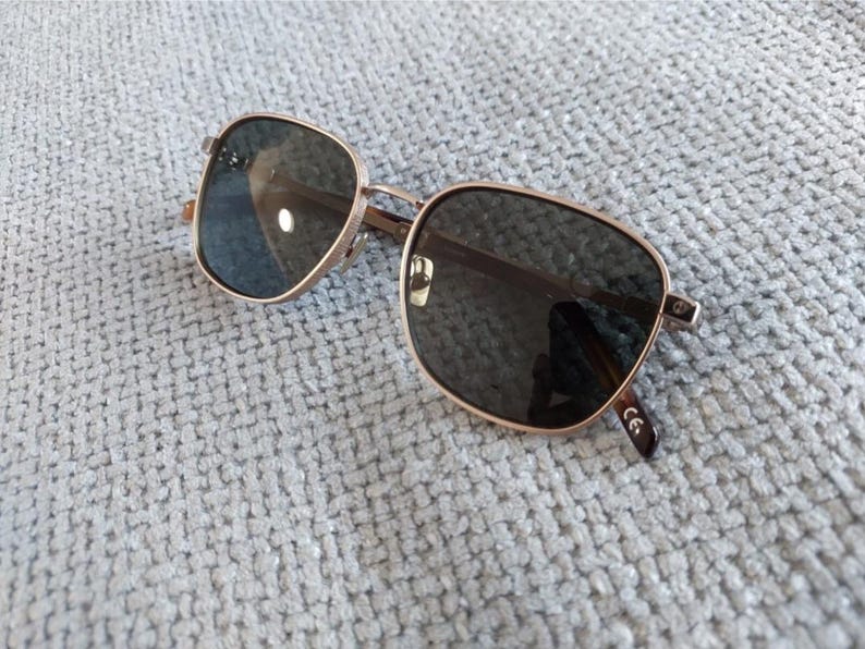 Puede incluir: Gafas de sol cuadradas con montura de metal dorado y lentes oscuras. Las gafas de sol est&aacute;n sobre una superficie texturizada de color gris claro. Las lentes son reflectantes y las patillas tienen un dise&ntilde;o sutil.