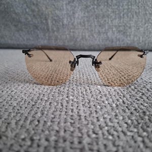 May include: Rimless sunglasses with a light brown tint. The lenses are oval-shaped and held together by a black bridge and temples. The glasses are resting on a gray, textured surface.