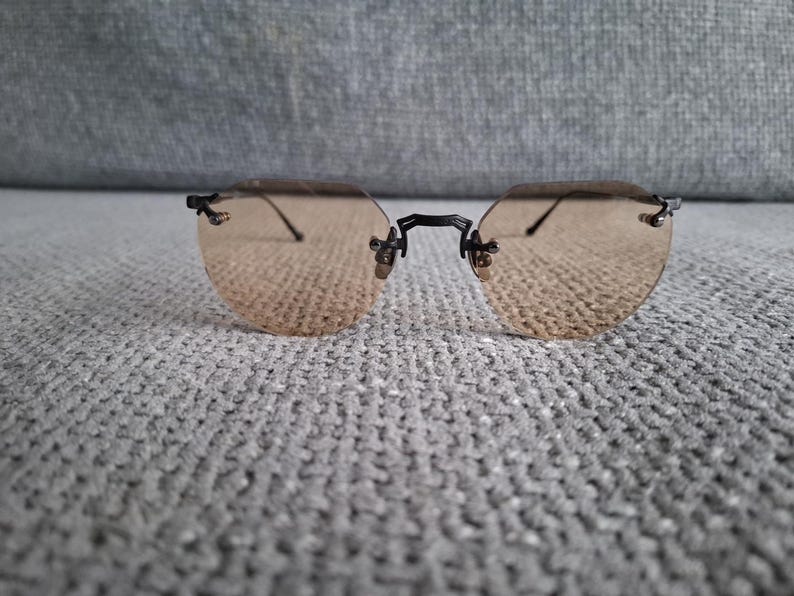 May include: Rimless sunglasses with a light brown tint. The lenses are oval-shaped and held together by a black bridge and temples. The glasses are resting on a gray, textured surface.
