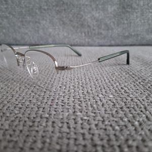 May include: A pair of eyeglasses with a silver-colored metal frame and clear lenses. The temples are a dark green color. The glasses are resting on a gray, textured surface.