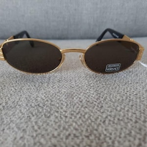 May include: Gold-coloured sunglasses with dark lenses. The oval lenses are set in a gold frame with black arms. A small white tag with the Versace logo is attached to the right lens.