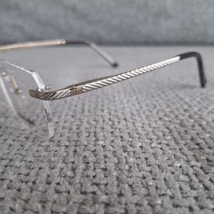 Op de afbeelding: Bril zonder montuur met heldere glazen en zilverkleurige monturen. De veren hebben een textuurpatroon en de ooreinden zijn zwart. De brug en scharnieren zijn goudkleurig. De bril ligt op een grijze achtergrond.