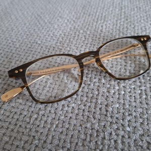 May include: Eyeglasses with a brown tortoiseshell frame and gold-coloured arms. The rectangular lenses are clear. The glasses are resting on a grey, textured surface.