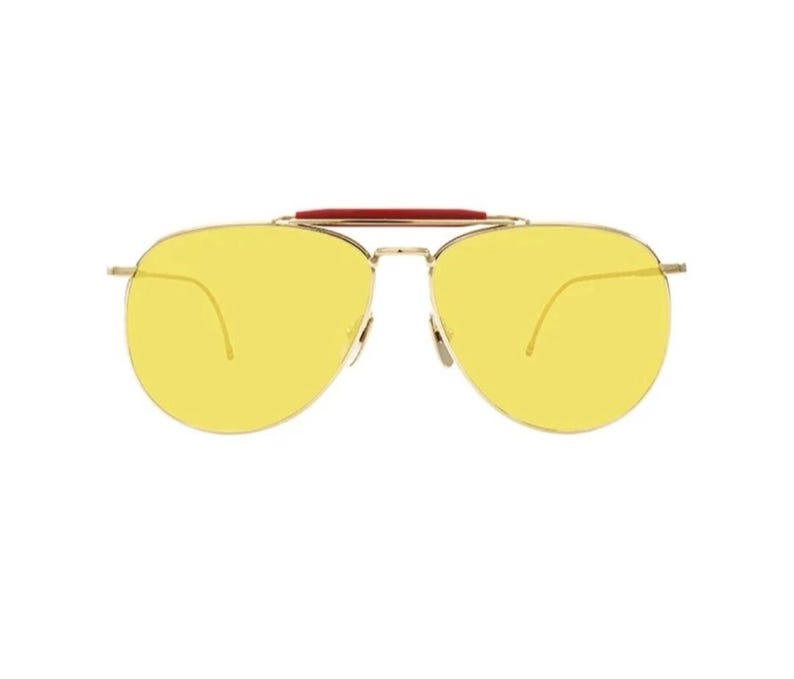 May include: Aviator-style sunglasses with a gold-colored frame and bright yellow lenses. The sunglasses feature a red bridge detail. The lenses are a vibrant yellow color, offering a bold and stylish look.