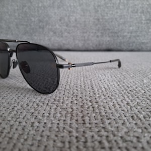 May include: Black aviator sunglasses with dark lenses and a double bridge. The sunglasses have a sleek, modern design with a thin frame and temples. The sunglasses are resting on a gray textured surface.