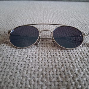 以下が含まれることがあります： ダークレンズのシルバーフレームサングラス。メガネはダブルブリッジとクラシックな丸い形をしています。テンプルは細く、耳の周りにカーブしています。サングラスは、グレーのテクスチャード加工された表面に置かれています。