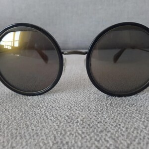 May include: Round sunglasses with black frames and dark lenses. The sunglasses have a thin metal bridge and temples. The lenses reflect a yellow hue. The sunglasses are resting on a textured, light gray surface.