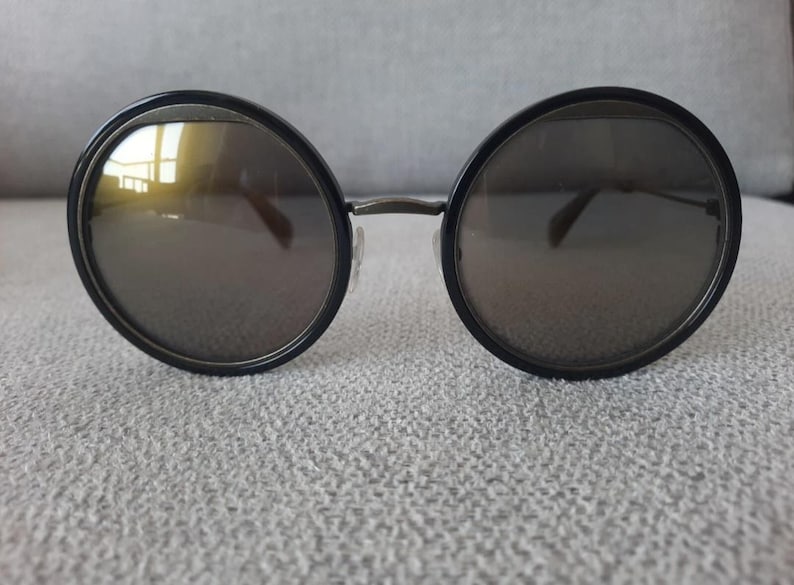 May include: Round sunglasses with black frames and dark lenses. The sunglasses have a thin metal bridge and temples. The lenses reflect a yellow hue. The sunglasses are resting on a textured, light gray surface.