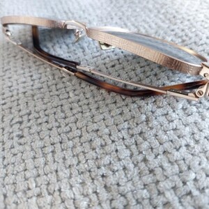Puede incluir: Gafas de color oro rosa con montura texturizada y lentes oscuras. Las patillas presentan un dise&ntilde;o de carey. Las gafas est&aacute;n parcialmente abiertas, apoyadas sobre una superficie texturizada de color gris claro.