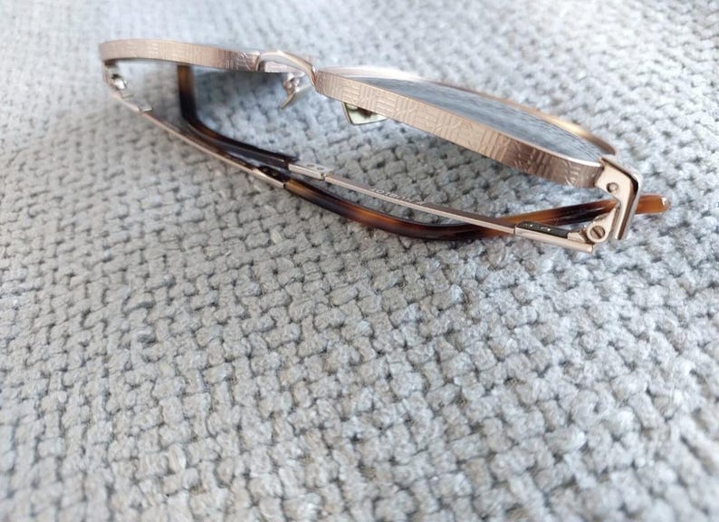 Puede incluir: Gafas de color oro rosa con montura texturizada y lentes oscuras. Las patillas presentan un dise&ntilde;o de carey. Las gafas est&aacute;n parcialmente abiertas, apoyadas sobre una superficie texturizada de color gris claro.