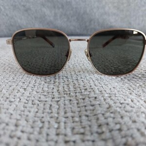 Puede incluir: Gafas de sol con montura dorada y lentes oscuras. Las gafas tienen forma cuadrada con bordes redondeados. El puente y las patillas son finos, y las lentes son de color gris oscuro. Las gafas est&aacute;n sobre una superficie texturizada de color gris claro.