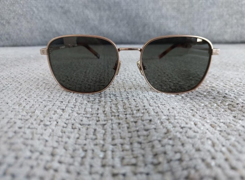 Puede incluir: Gafas de sol con montura dorada y lentes oscuras. Las gafas tienen forma cuadrada con bordes redondeados. El puente y las patillas son finos, y las lentes son de color gris oscuro. Las gafas est&aacute;n sobre una superficie texturizada de color gris claro.