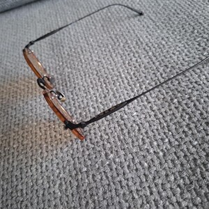 May include: Eyeglasses with a thin, black metal frame and amber-tinted lenses. The glasses are resting on a gray, textured surface. The temples of the glasses are thin and extend outward.
