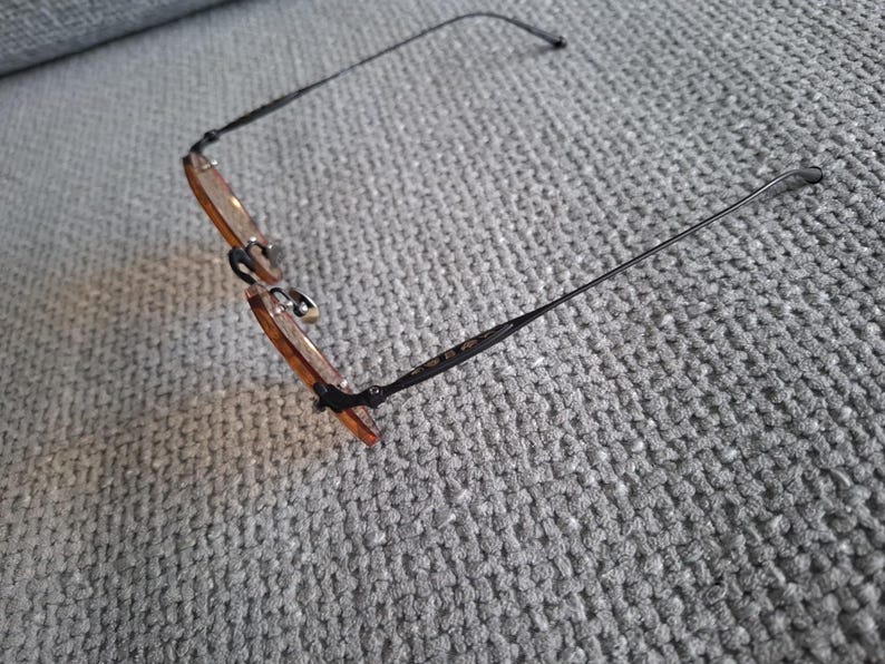 May include: Eyeglasses with a thin, black metal frame and amber-tinted lenses. The glasses are resting on a gray, textured surface. The temples of the glasses are thin and extend outward.
