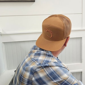 May include: A brown and tan baseball cap with a mesh back. The cap has a brown embroidered logo of a fish hook on the front.