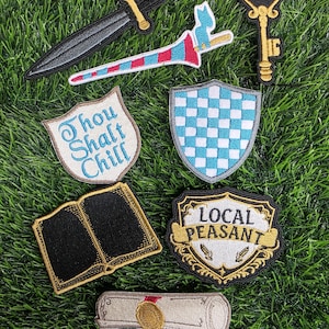 May include: Assortment of embroidered patches on green grass. Includes a black dagger, a red and blue lance, a gold key, two shields, an open book, a scroll, and a patch that says "Local Peasant".