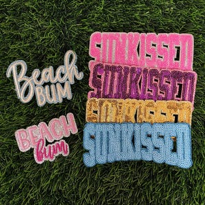 May include: A collection of sequined patches with the words "Sunkissed" and "Beach Bum". The "Sunkissed" patches are in pink, purple, gold, and blue. The "Beach Bum" patches are in pink and blue.