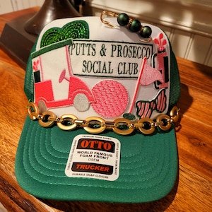May include: A green trucker hat with a white patch that says "Putts & Prosecco Social Club". The patch features a pink golf cart with a golf flag and a golf ball. The hat has a gold chain around the brim and a small gold safety pin with green beads.