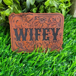 May include: A brown leather patch with the word "WIFFY" in black, surrounded by floral and scrollwork designs. The patch is rectangular with rounded corners. The background is green.