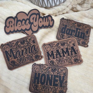 May include: Five brown leather patches with black lettering and floral designs. The patches read "Bless Your", "darlin'," "Mama", "MAMA", and "HONEY".