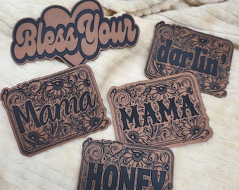 Könnte beinhalten: Fünf braune Leder-Patches mit schwarzer Beschriftung und floralen Designs. Die Patches lauten "Bless Your", "darlin'," "Mama", "MAMA" und "HONEY".