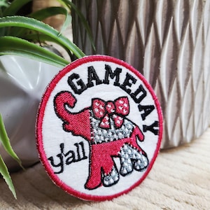 May include: A round, white patch with a red border featuring a red elephant design with a bow tie and the words "GAMEDAY y'all" in black text. The elephant has a silver and white pattern.