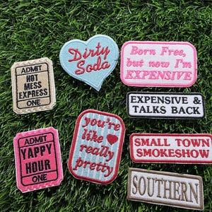 May include: A collection of embroidered patches with various phrases and designs. The patches include text like "Dirty Soda," "Born Free, but now I'm EXPENSIVE," and "You're like really pretty." The patches are in various shapes and colors.