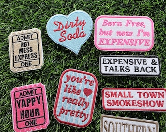 Può includere: Una collezione di toppe ricamate con varie frasi e disegni. Le toppe includono testo come "Dirty Soda", "Born Free, but now I'm EXPENSIVE" e "You're like really pretty". Le toppe sono di varie forme e colori.