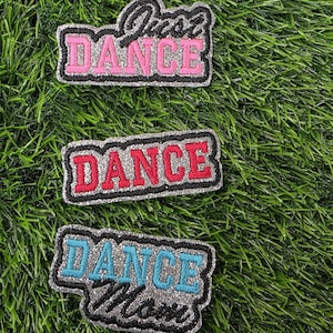 May include: Three glittery patches with the word "DANCE" in different colors: pink, red, and blue. One patch says "Just Dance" in black script. All patches have a black border and are on a green, grassy background.