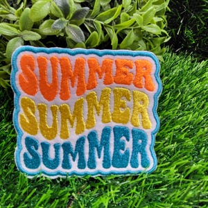 May include: A colorful patch with the word "SUMMER" in orange, yellow, and teal, stacked three times. The patch has a white background and a light blue border. The patch is on a bed of green grass.