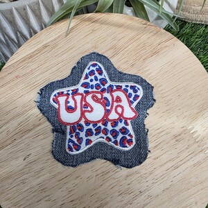 Pu&ograve; includere: Toppa a forma di stella in denim con la scritta "USA" ricamata in bianco con contorni rossi. L'interno della stella presenta un motivo leopardato blu e rosso. La toppa &egrave; probabilmente per abbigliamento o accessori.