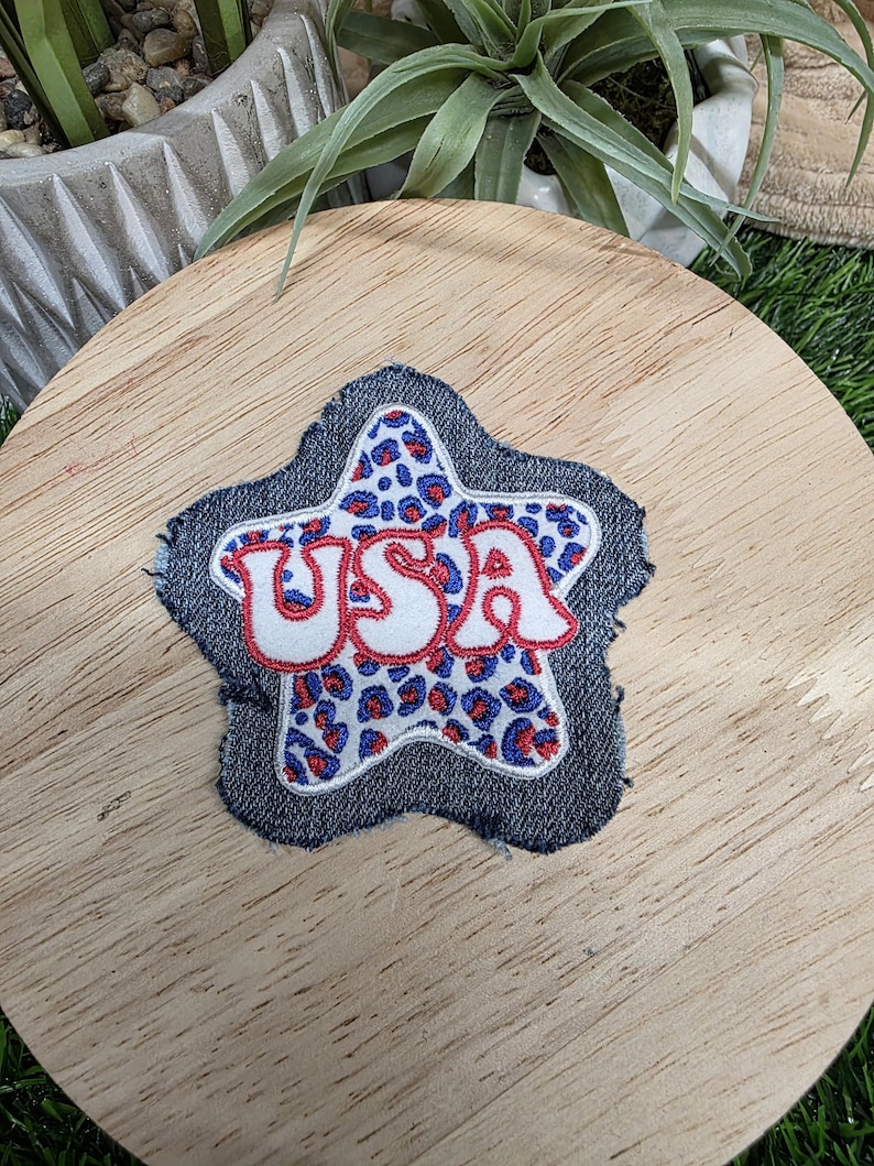 Pu&ograve; includere: Toppa a forma di stella in denim con la scritta "USA" ricamata in bianco con contorni rossi. L'interno della stella presenta un motivo leopardato blu e rosso. La toppa &egrave; probabilmente per abbigliamento o accessori.