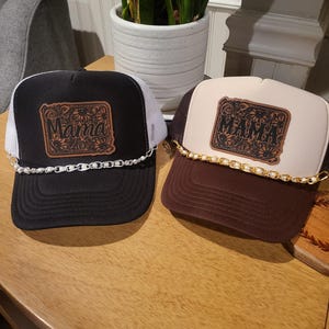 May include: Two trucker hats with a brown leather patch that says "Mama" in a decorative font. The hats are black and brown with white mesh back panels. Both hats have a gold chain around the brim.